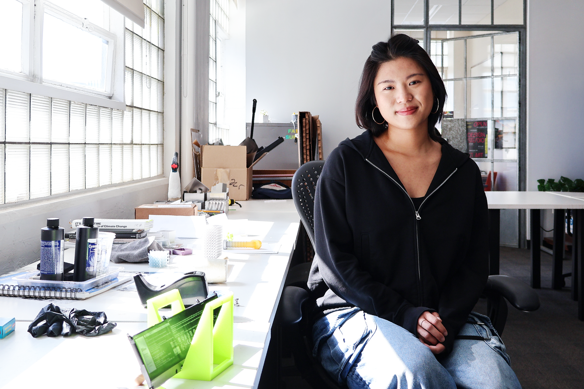 Young woman seated at a bright work desk in a studio or office space, smiling at the camera. She is wearing a black zip-up hoodie and blue jeans. The desk is scattered with design tools, notebooks, and prototypes, with natural light streaming in from glass block windows.